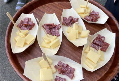 A wooden tray with six small bowls, filled with pieces of cheese and sausage. The bites are attached to wooden sticks.