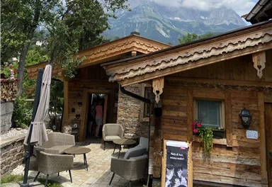 A cozy wooden house in the mountains with terrace furniture and trees. In the background, there are large mountains and a cloudy sky.