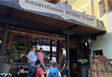 A farm shop named "Wilder Käser" can be seen. A few people are standing in front of the entrance and are having a conversation.