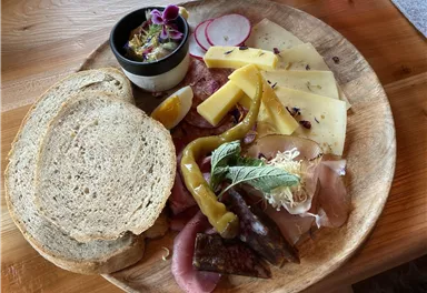 A rustic platter with various types of cheese, sausage, bread, and fresh vegetables. There is also a small dip in a bowl.