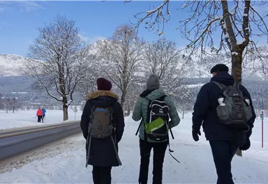 Drei Personen wandern auf einem schneebedeckten Weg. Im Hintergrund sind schneebedeckte Berge und Bäume zu sehen.