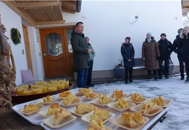 A group of people is standing in front of a building and listening to a speaker. On a table in front of them, several plates of food are arranged.