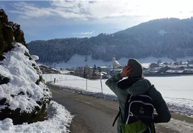 A person stands on a snowy road and looks at the mountains. In the background, wooden buildings and a church tower can be seen.