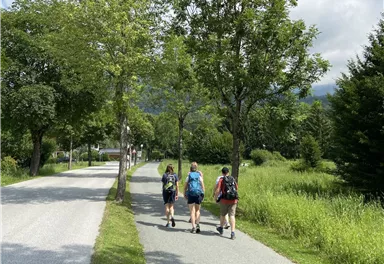 Three hikers are walking on a narrow path lined with trees. The surroundings are green and invite exploration.