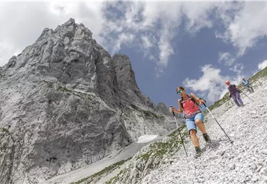 Eine Gruppe von Wanderern erklimmt einen steilen, steinigen Hang in den Bergen. Im Hintergrund ist ein imposanter Felsen unter einem bewölkten Himmel zu sehen.