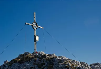 Ein großes Kreuz steht auf einem Berggipfel unter einem klaren blauen Himmel. Die umliegenden Felsen sind schroff und von geringer Vegetation geprägt.