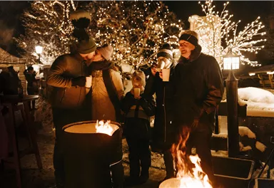 Eine gemütliche Winteratmosphäre mit Menschen, die um ein Feuer stehen und heiße Getränke genießen. Die Umgebung ist festlich beleuchtet mit Weihnachtslichtern.