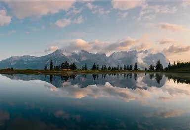 A tranquil mountain landscape with reflective water and high mountains in the background. The sky is interspersed with soft clouds and warm colors.