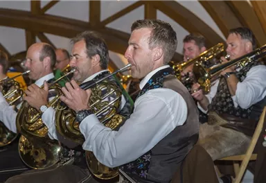 A group of musicians plays horns in traditional attire. They are in a festive room with wooden beams.