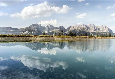 A tranquil landscape with a clear lake and majestic mountains in the background. The mountains are perfectly reflected in the water.