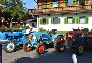 Drei bunte Traktoren stehen vor einem traditionellen bayerischen Gebäude. Der Himmel ist klar und die Umgebung ist grün und einladend.