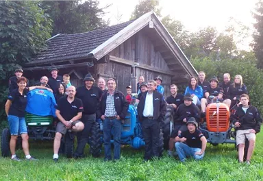 Eine Gruppe von Menschen steht vor einer alten Scheune mit Traktoren. Alle tragen schwarze T-Shirts und lächeln in die Kamera.