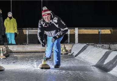 A man is playing curling on a lit track. In the background, spectators in winter clothing can be seen.
