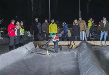 A group of people stands at the edge of an ice rink while a child plays on the ice with a stick. It is a cold winter night and the spectators are dressed warmly.