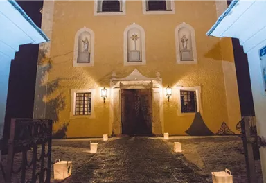 A pretty building with a yellow facade and illuminated windows stands in the dark. Several glowing lanterns are placed at the entrance.