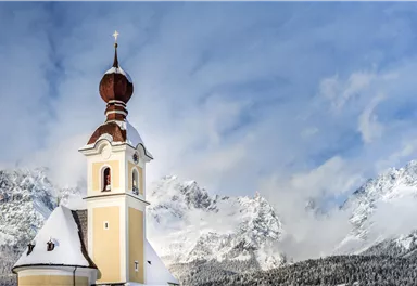 Eine hübsche Kirche mit einer charakteristischen Kuppel steht im Schnee. Im Hintergrund sind majestätische Berge und ein blauer Himmel zu sehen.