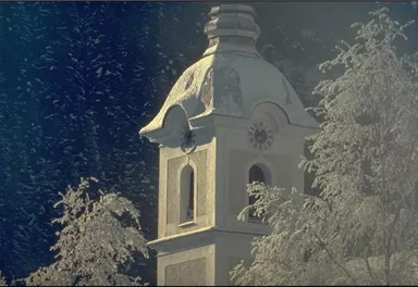 A snowy church tower with an elegant roof stands between snow-covered trees. In the background, a clear winter sky can be seen.