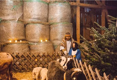 A traditional nativity scene with Mary and Joseph, surrounded by animals. In the background, there are hay bales and a Christmas chimney.