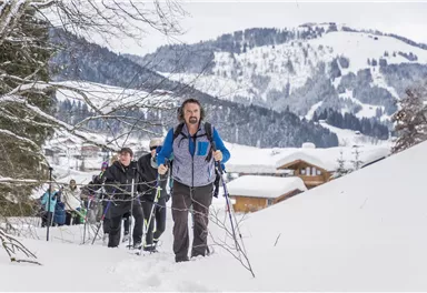 A group of hikers is climbing a snow-covered slope in the mountains. The surroundings are wintry with snow-covered trees and cabins in the background.