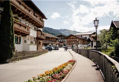 Eine malerische Straße in einer Alpenstadt mit traditionellen Holzhäusern und bunten Blumenbeeten. Im Hintergrund sind Berge und ein blauer Himmel zu sehen.