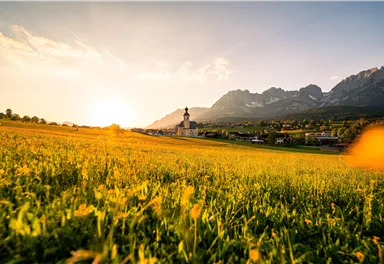 Eine malerische Landschaft mit einer Wiese voller Blumen und majestätischen Bergen im Hintergrund. Die Sonne geht hinter der Kirche unter und schafft eine warme Atmosphäre.