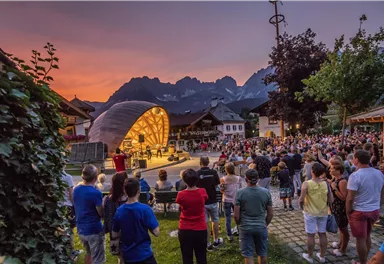 Ein Konzert im Freien bei Sonnenuntergang mit einer großen Menschenmenge. Die malerische Berglandschaft bildet eine beeindruckende Kulisse.