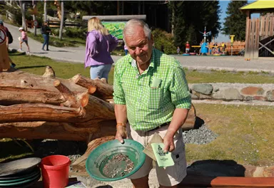 An older man stands at the table and shows a shovel with gold mining equipment. In the background, playground equipment and trees are visible.
