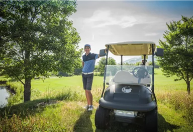 A man is standing next to a golf cart on a golf course. In the background, there are trees and a green meadow visible.