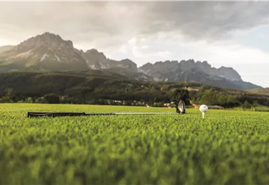 A golf course with green grass and a golf ball on a tee. In the background, impressive mountains and a cloudy sky can be seen.