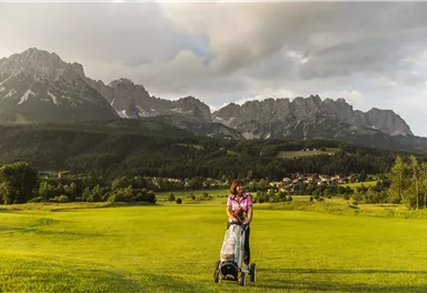 Eine Frau mit einem Golfwagen steht auf einem grünen Golfplatz. Im Hintergrund sind beeindruckende Berge und ein bewölkter Himmel zu sehen.