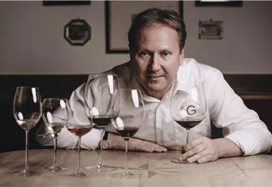 A man is sitting at a table and holding a wine glass in his hand. In front of him are five wine glasses with different types of wine.