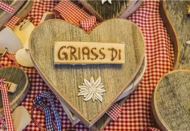 A wooden heart with the inscription "GRIASS DI" and a decorative flower. The background is decorated with checked fabric.