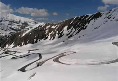 A picturesque mountain landscape with snow-covered hills and winding roads. The sky is clear and blue, offering a breathtaking view.