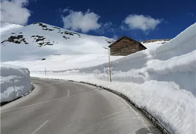A snow-covered road surrounded by high snow banks. In the background stands a small wooden house under a blue sky with some clouds.