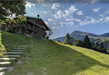 A picturesque country house on a green meadow with gentle hills in the background. The sky is clear and blue, with a few clouds.