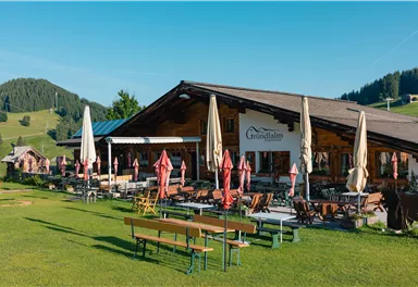 Ein gemütliches Restaurant in der Alpenlandschaft mit einer großen Terrasse. Umgeben von grünen Wiesen und bewaldeten Hügeln.