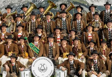 A large group of musicians in traditional costumes stands in front of a mountain landscape. They hold various instruments and smile at the camera.