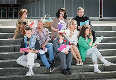 Eine Gruppe von Personen sitzt auf Stufen und hält verschiedene Dokumente in den Händen. Sie scheinen eine Aufführung oder ein Casting zu proben.