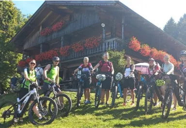 A group of cyclists is smiling on a meadow in front of a traditional wooden house. In the background, trees and blooming plants can be seen.