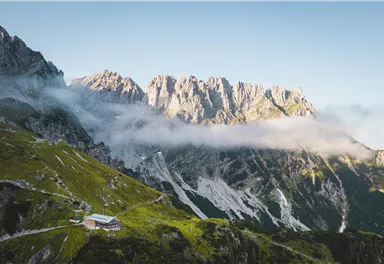 An impressive mountain landscape with high peaks and cloud-covered slopes. In the foreground, a cozy cabin can be seen.