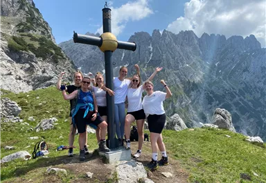 A group of people stands in front of a mountain cross in the mountains. They are smiling and happily posing for the photo.