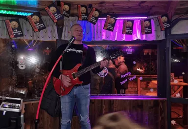 Ein Musiker mit einer roten Gitarre steht auf einer kleinen Bühne und singt. Im Hintergrund sind bunte Lichter und festliche Dekorationen zu sehen.