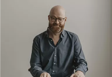 Ein Mann mit Brille sitzt an einem Keyboard. Er trägt ein blaues Hemd und lächelt freundlich.