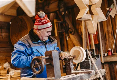 Ein älterer Mann arbeitet in einer Holzwerkstatt. Er dreht ein Stück Holz auf einer Drehmaschine und trägt eine blaue Jacke und eine Mütze.