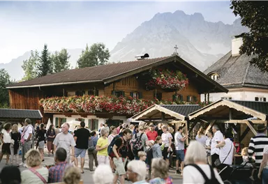 Ein belebter Marktplatz mit vielen Menschen vor einem traditionellen Holzhaus. Im Hintergrund sind Berge und Bäume zu sehen, die eine idyllische Atmosphäre schaffen.