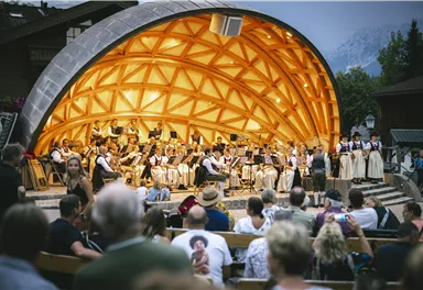 Ein Orchester spielt in einem einzigartigen Pavillon vor einem Publikum. Menschen sitzen auf Bänken und genießen das Konzert.