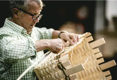 Ein älterer Mann mit Brille arbeitet an einem Holzstuhl. Er ist konzentriert und verwendet verschiedene Techniken zur Fertigung.