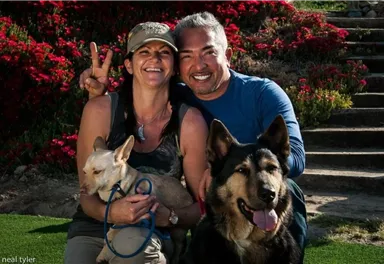 A joyful couple is sitting outdoors, holding two dogs in their hands. Colorful plants are blooming in the background.
