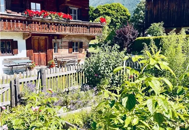 Ein traditionelles Holzhaus mit Blumenkästen und einem schönen Garten. Im Hintergrund sind Berge und ein blauer Himmel zu sehen.