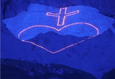 Eine herzförmige Lichtinstallation mit einem Kreuz ist auf einer schneebedeckten Fläche sichtbar. Das Bild hat eine mystische blaue Beleuchtung.